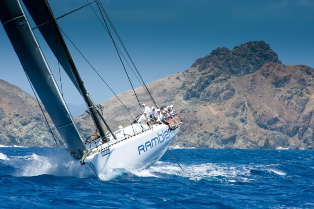 A sailing boat named Rambler racing on the ocean with a mountainous coastline in the background