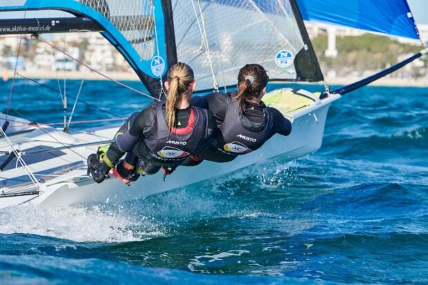Two sailors hanging off the side of a sailboat racing on vibrant blue water