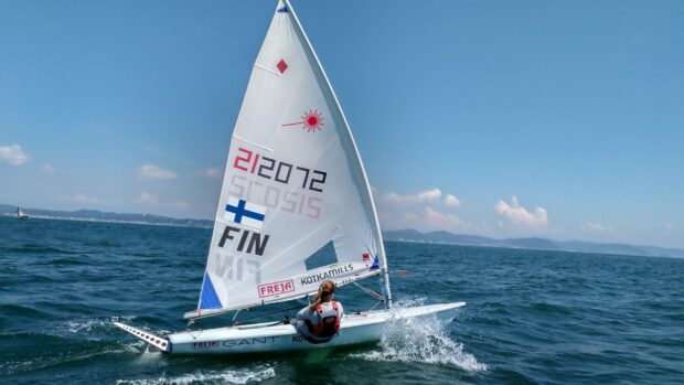 A sailor navigating a small sailboat on open water with clear blue sky and distant mountains