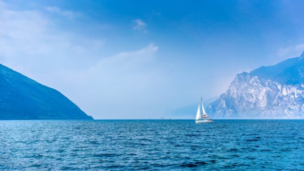 A sailing boat glides across the calm blue sea with mountains in the background