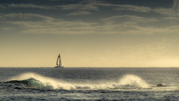 A sailing boat moving across the ocean waves under a cloudy sky at sunset
