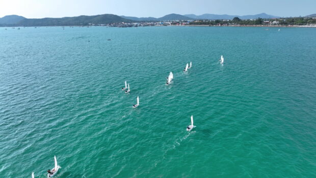 A fleet of sailboats racing on clear turquoise water near a coastal town with mountains in the background