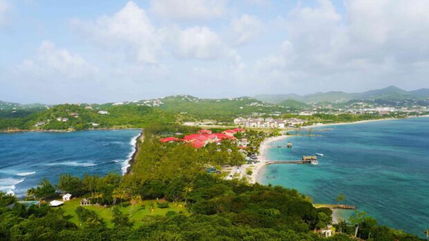 A lush green landscape of Saint Lucia with red roofed buildings and turquoise ocean bays under a cloudy sky