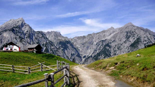 A scenic mountain path runs through grassy fields with alpine houses and rocky peaks in Austria, 4K Desktop Wallpaper