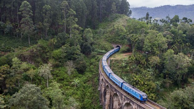 A blue train travels over a stone bridge surrounded by green forest in Sri Lanka, 2K Desktop Wallpaper