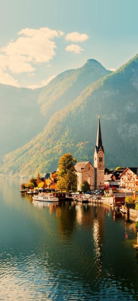 A picturesque village with a church and boats by the calm lake mountains in Austria, 2K Mobile Wallpaper