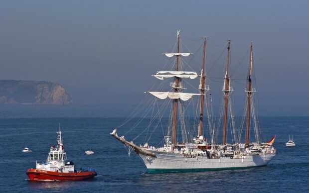 A schooner sailing on the ocean near a coastline with a tugboat beside it