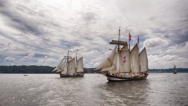 A schooner sailing on calm water under a cloudy sky with a forest in the background