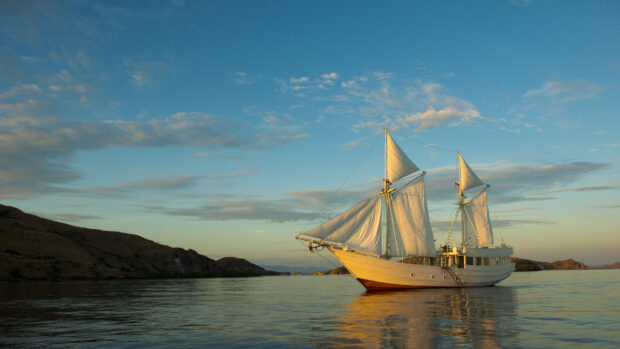 White schooner sailing near rocky hills under a blue sky at sunset
