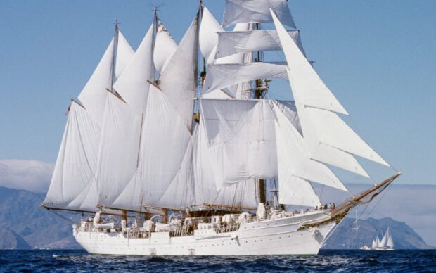 A classic schooner sailing on calm blue waters with multiple white sails fully unfurled