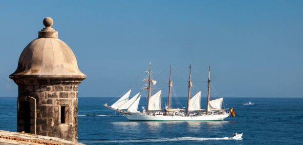 A schooner sailing on the ocean near an old stone tower with a clear blue sky