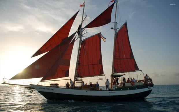 A classic schooner with red sails sailing on the ocean at sunset with people on board