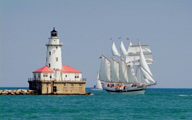 A schooner sailing near a lighthouse on a clear blue sea with bright skies