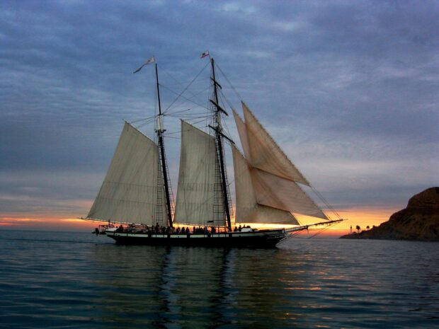 A schooner sailing on calm water during sunset with hills and palm trees in the background