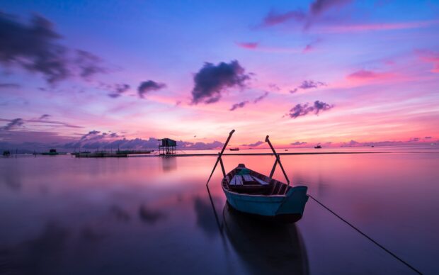 A small boat rests on calm water during a vibrant sunset with colorful clouds, 2K Desktop Wallpaper