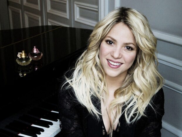 Shakira with blonde hair smiling near a piano in a black outfit, HD Desktop Wallpaper