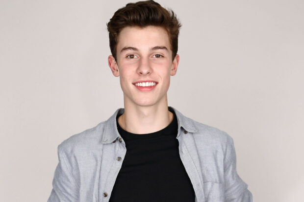 Shawn Mendes posing with a friendly smile wearing a gray shirt and black t shirt against a neutral background