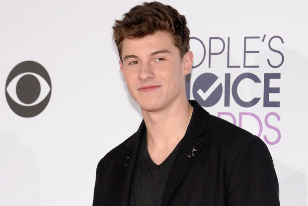 Shawn Mendes smiling in a black jacket at an award event with a white background