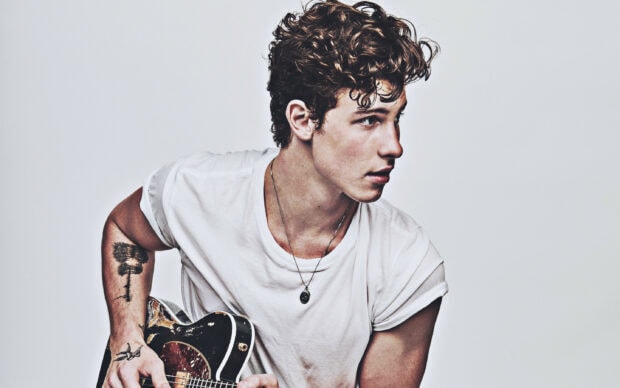 Shawn Mendes wearing a white t shirt playing a guitar with tattoos on his arm