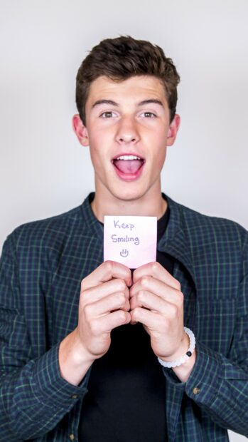 Shawn Mendes holding a note with keep smiling text and wearing a checkered shirt