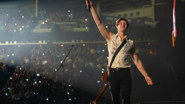 Shawn Mendes raising his hand with a guitar on stage during a live concert with audience lights in the background