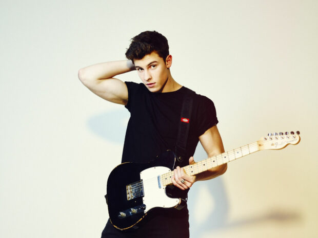 Shawn Mendes holding a guitar wearing a black shirt in a studio setting