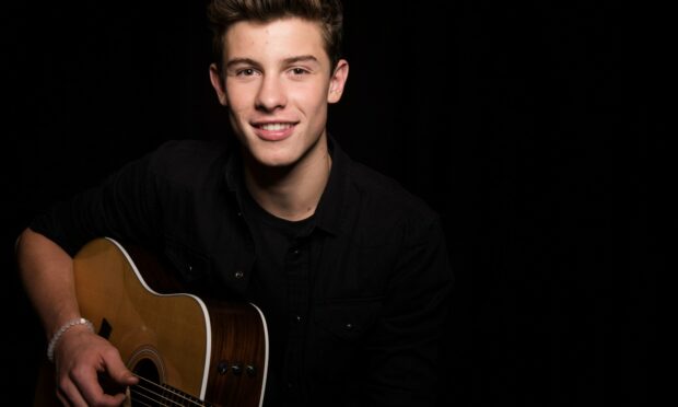 Shawn Mendes playing guitar with a smile in a dark setting