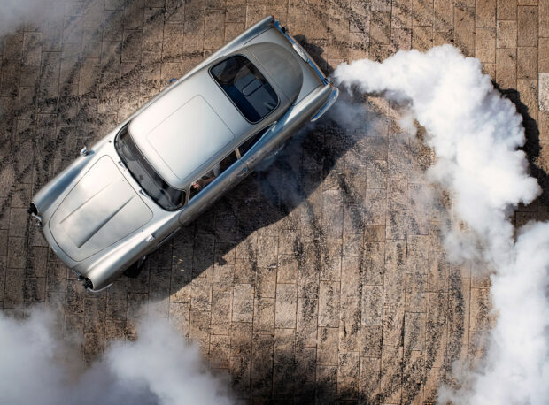 A silver sports car is drifting leaving smoke trails on a tiled surface in this No Time To Die scene, HD Desktop Wallpaper
