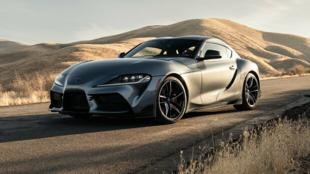 Sports car, Toyota Supra, silver coupe on scenic mountain road with dry grass and rolling hills in the background, luxury vehicle, daytime outdoor setting, high-performance automobile.