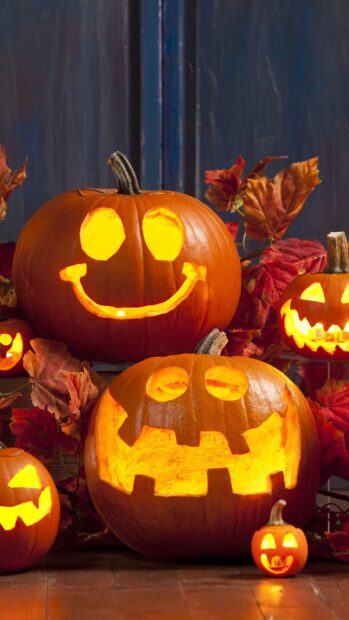A group of glowing Halloween pumpkins with carved faces sits among autumn leaves on a wooden floor, 2K Mobile Wallpaper