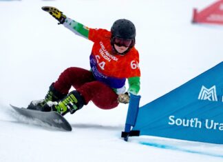A snowboarder wearing red pants and a green sleeve jacket racing past a gate in a snowboarding competition