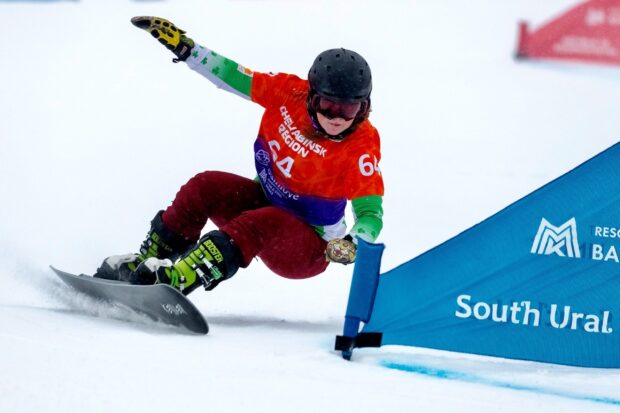 A snowboarder wearing red pants and a green sleeve jacket racing past a gate in a snowboarding competition