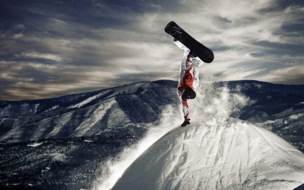Snowboarding athlete performing a handstand trick on a snowy slope with mountains in the background