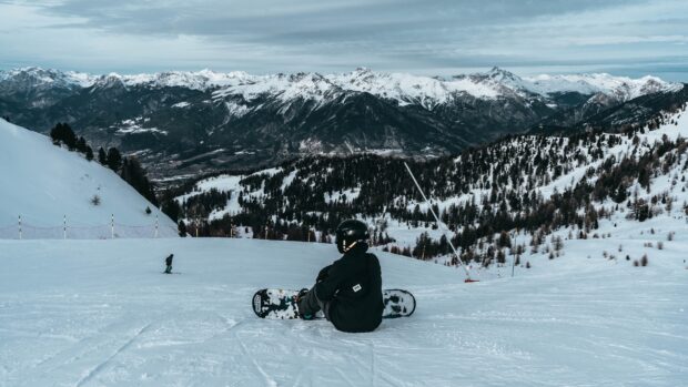 A snowboarder sitting on snow with snowboarding gear and mountains in the background
