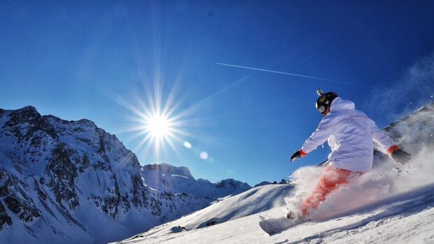 A snowboarder wearing white jacket and red pants sliding down the snowy mountain slope in bright sunlight
