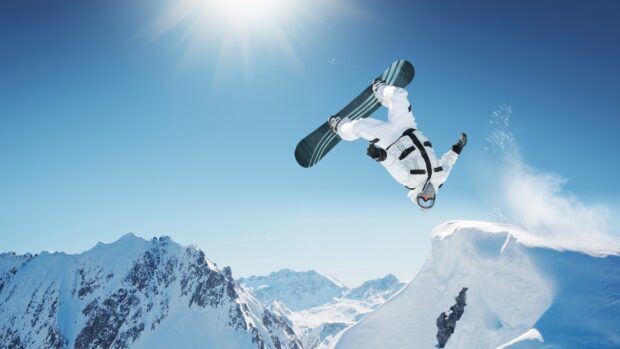 A snowboarder performing a flip in midair over snowy mountains under a clear blue sky