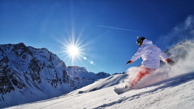 A snowboarder dressed in white and orange snowboarding on a sunny snowy mountain slope
