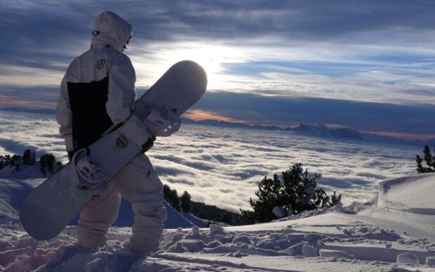 A snowboarder standing on snowy mountain top overlooking clouds and distant peaks