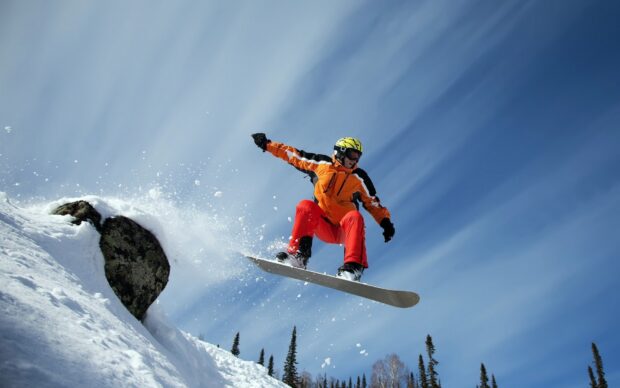 A snowboarder wearing orange gear is jumping over snow covered terrain on a bright winter day