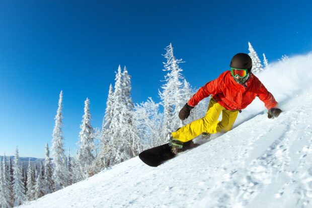 A snowboarder wearing yellow pants and a red jacket riding down a snowy slope surrounded by snowboarding