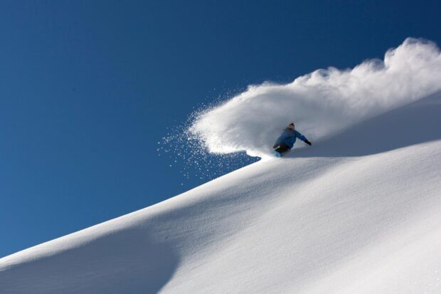 Snowboarder carving through fresh powder on a snowy mountain slope under clear blue sky