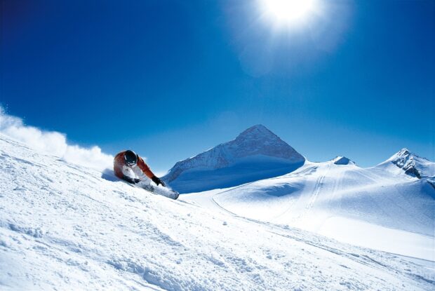 A snowboarder carving down a snowy slope with mountains in the background on a sunny day