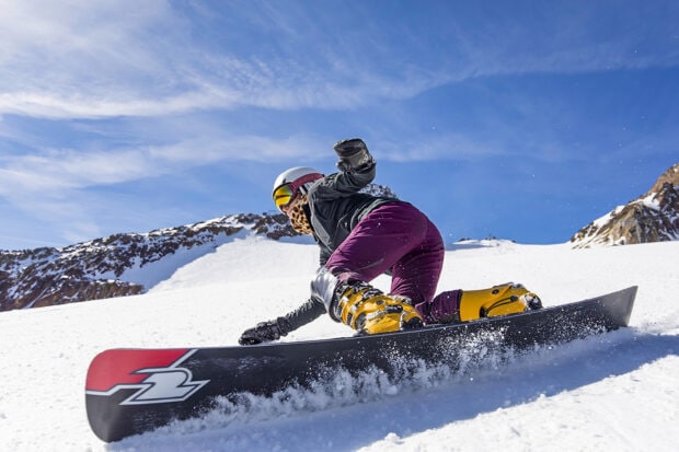 A snowboarder wearing yellow boots and purple pants carving on a snowboarding slope