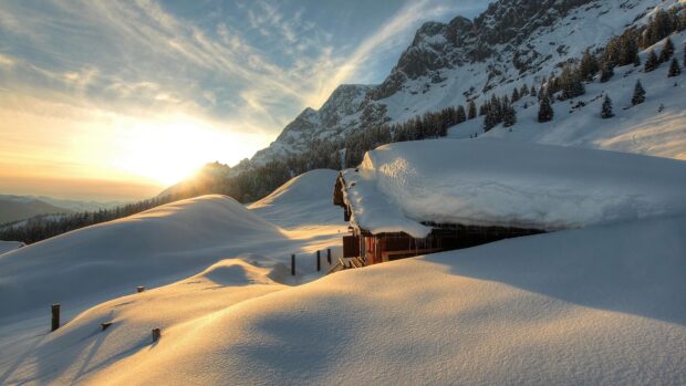 A snowy cabin is nestled among snow covered hills with the sun setting behind the mountains in Austria, 4K Desktop Wallpaper