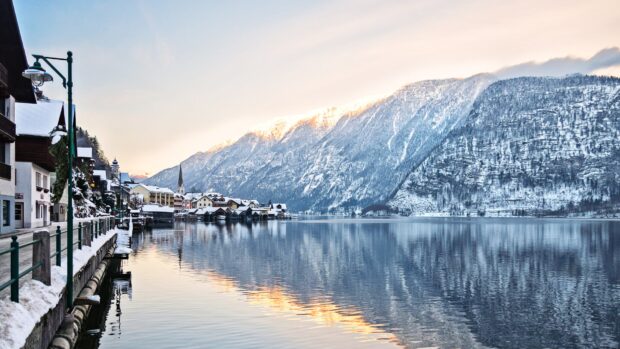 A snowy village sits beside a calm lake reflecting towering mountains in Austria, 4K Desktop Wallpaper