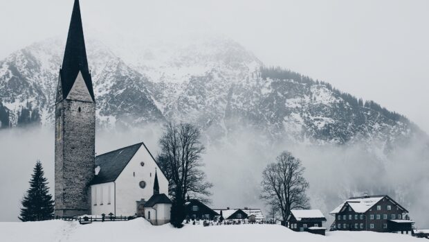 A snowy village scene in Austria with a church and houses covered in snow and misty mountains behind, 4K Desktop Wallpaper