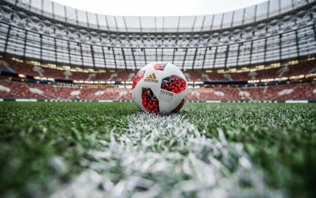 A soccer ball on the field at the center line in a large stadium with fans in the background