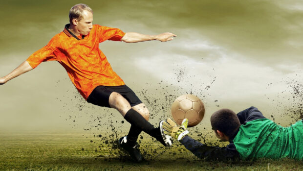 Soccer player kicking the ball while the goalkeeper dives to save the ball on the field