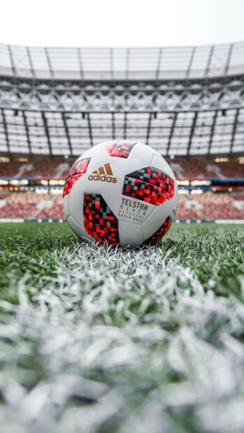 A soccer ball with red and black patterns resting on the grass at the center line of a stadium