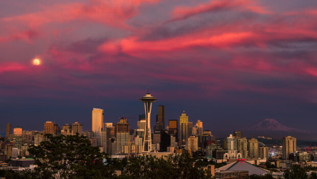 The Space Needle stands tall over the Seattle skyline at sunset with a colorful sky and a visible moon